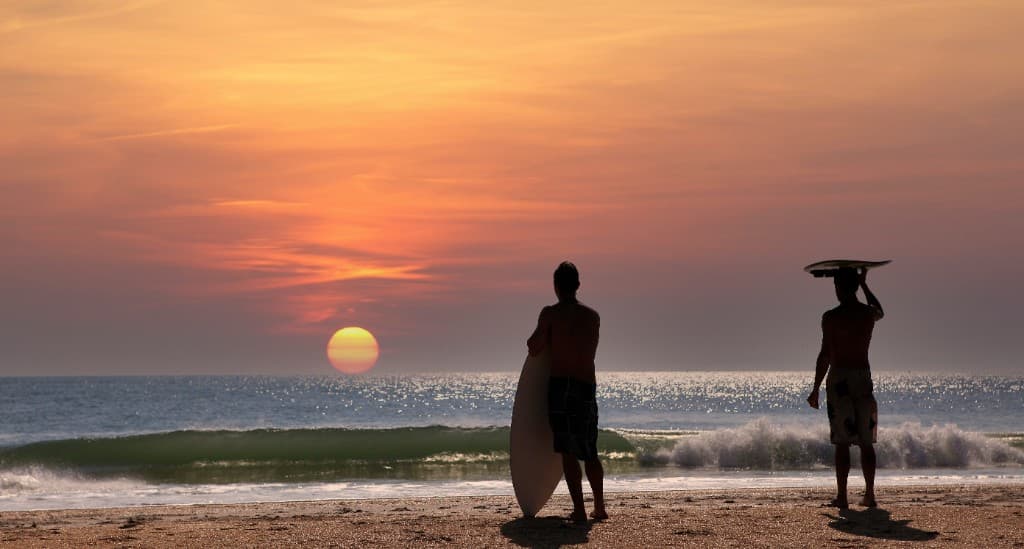 Surfers standing on Cocoa Beach at sunset with ocean waves and a warm orange sky.